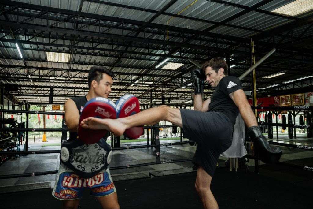An Asian Muay Thai trainer holds red pads while a Caucasian fighter executes a high kick during a sparring session in a spacious indoor gym.