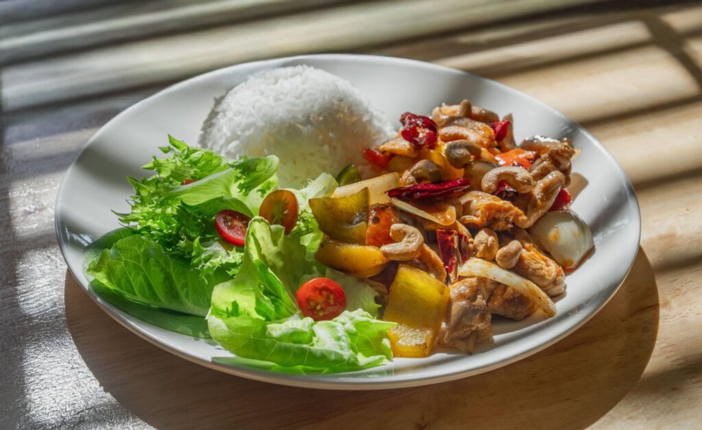 A plate of white rice with spicy stir-fried chicken, cashews, vegetables, and a fresh green salad sits in bright natural light.