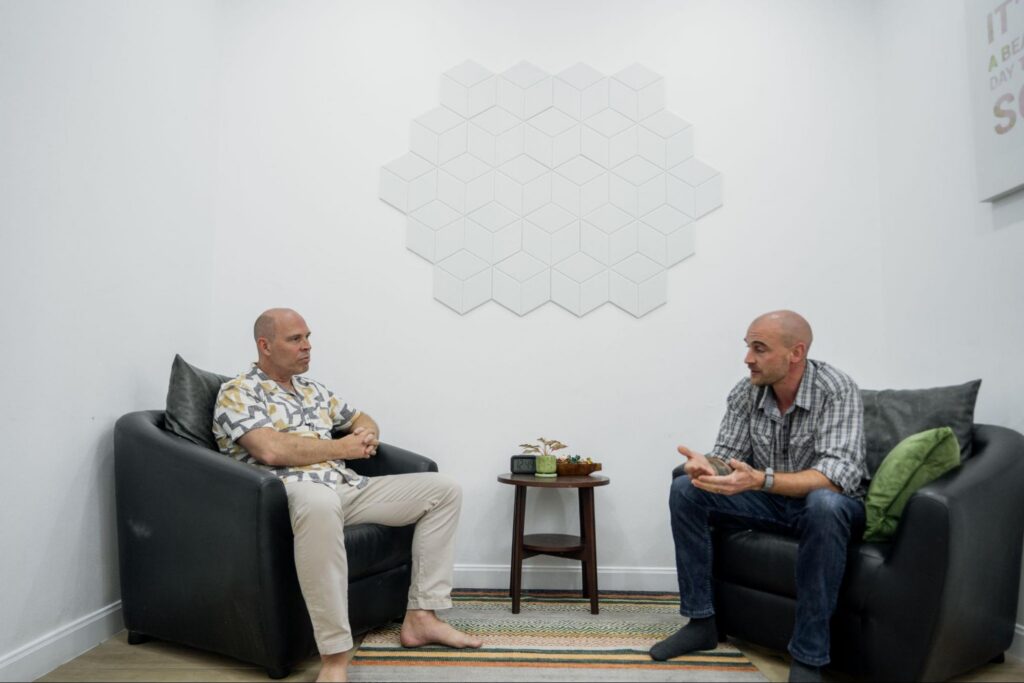 Two men seated in armchairs during a one-on-one counselling session, engaged in conversation in a calm, minimalist therapy room.